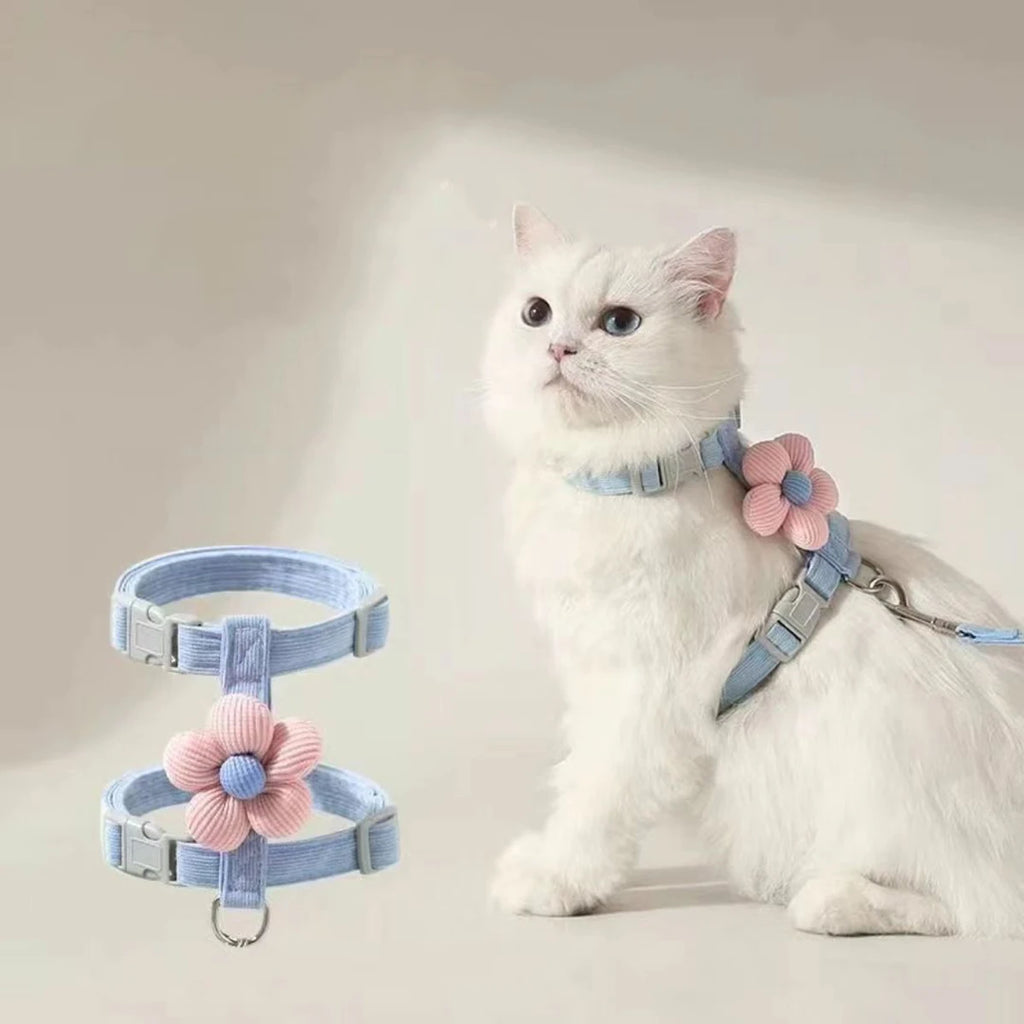 White cat wearing a light blue L-shape harness with pink floral attachments on a plain background