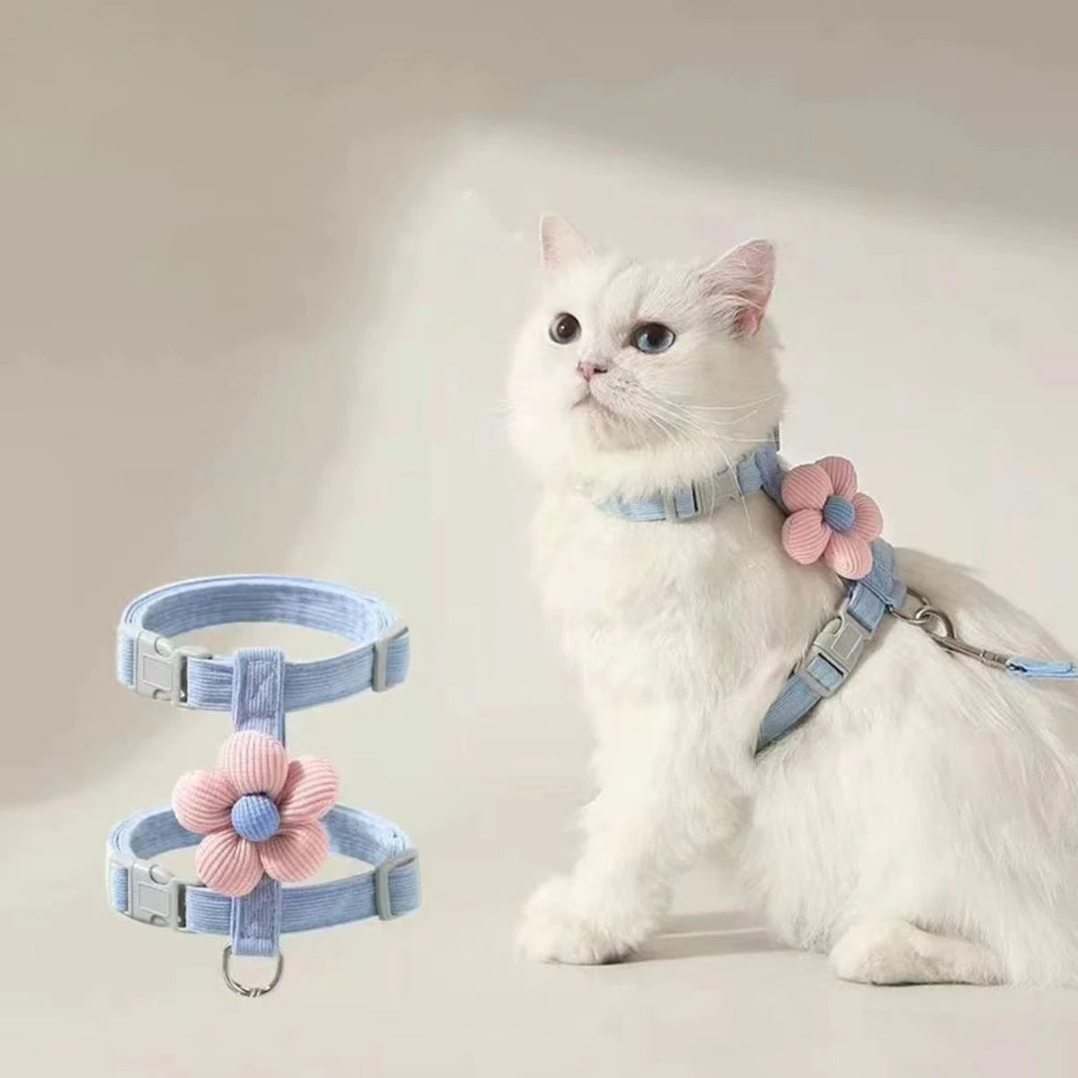 White cat wearing a light blue L-shape harness with pink floral attachments on a plain background
