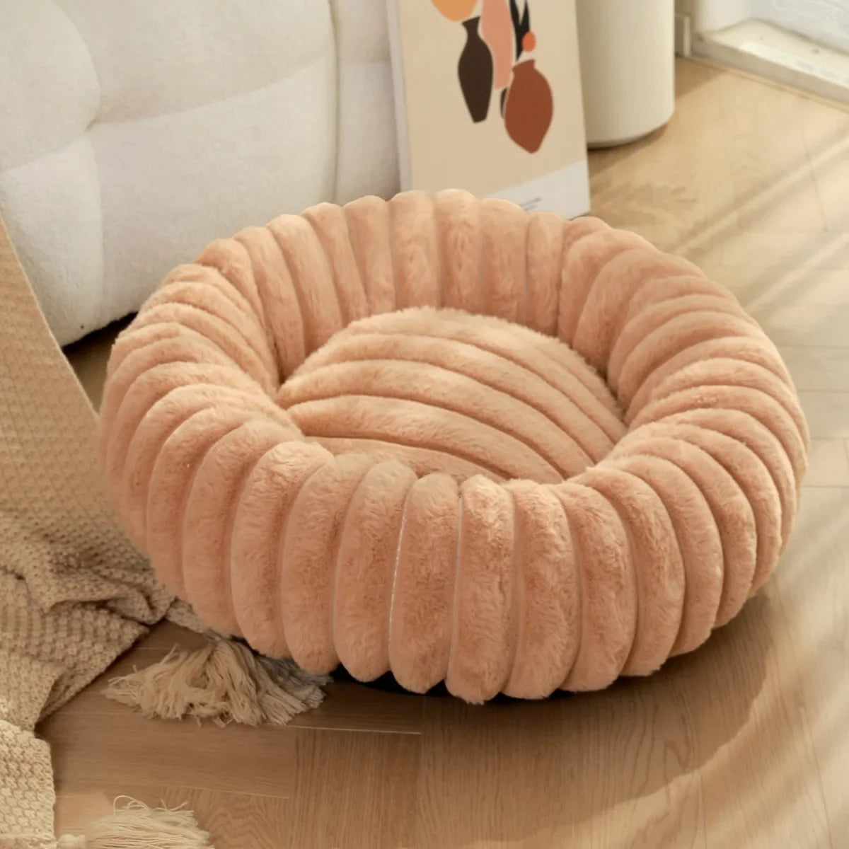 Peach Round, plush pet bed on a wooden floor with a couch and decorative pillow in the background.