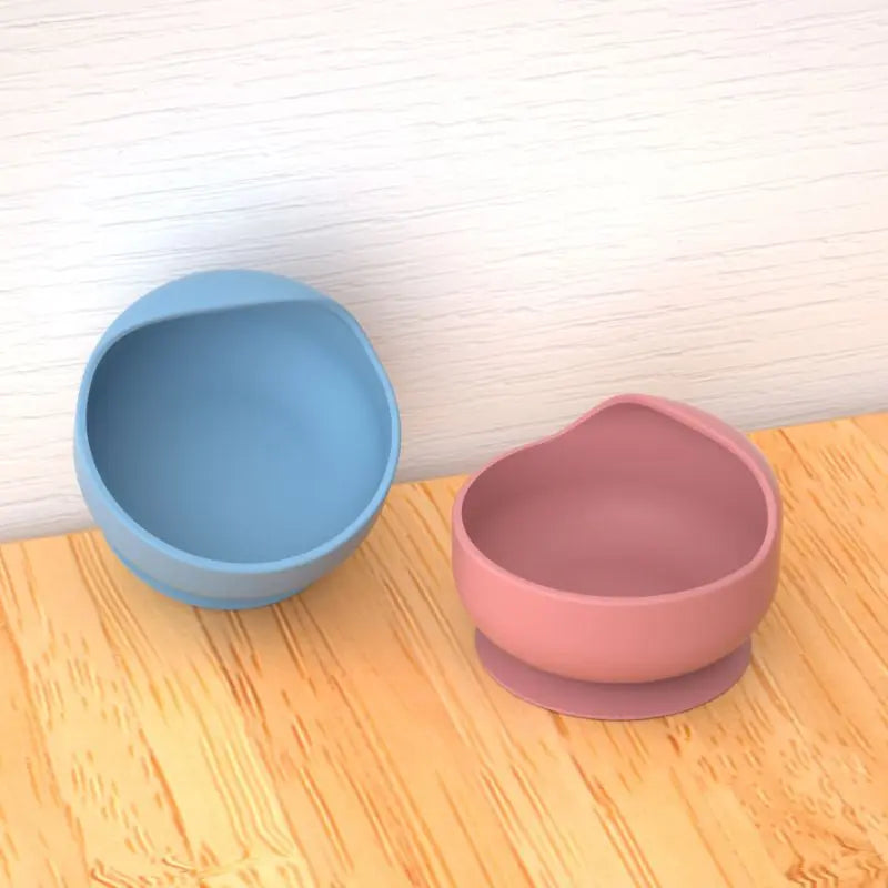 Two silicone bowls, one blue and one pink, on a wooden surface with a white wall background.
