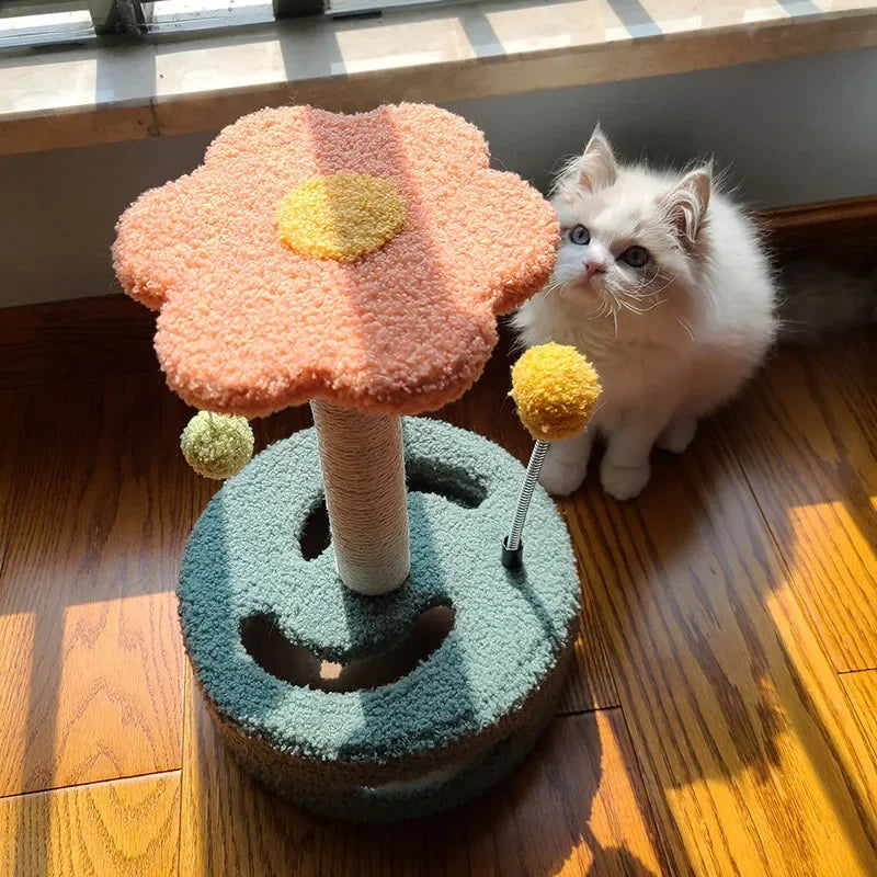 A fluffy white kitten sitting beside a flower-shaped cat scratching post with toys attached.