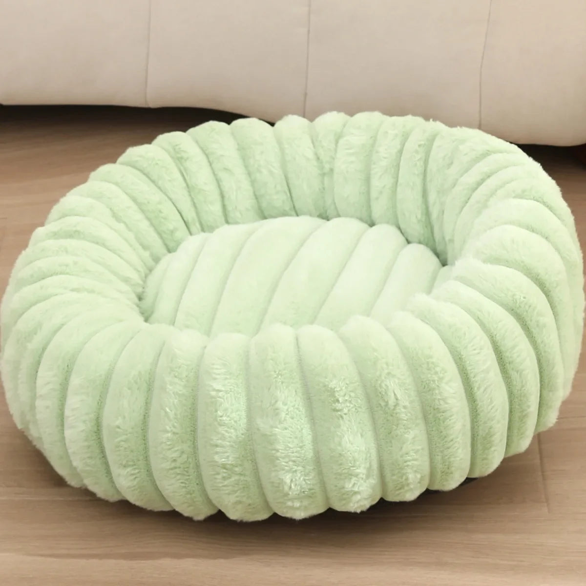 Light green plush pet bed on a wooden floor with a beige couch in the background