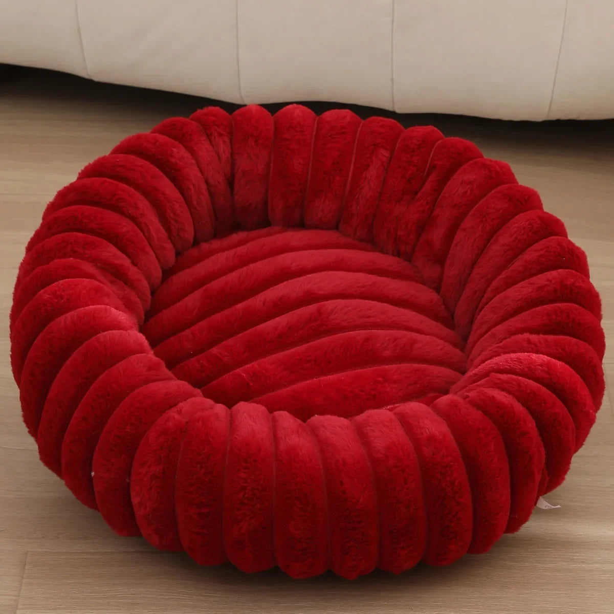 Red plush pet bed on a wooden floor with a beige couch in the background