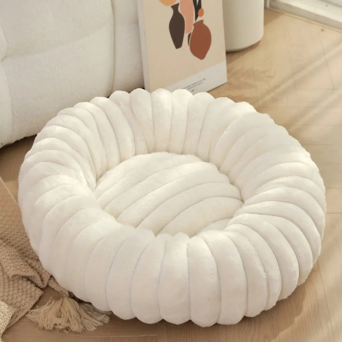 White round, plush white cushion on a wooden floor with a beige sofa and decorative pillow in the background.