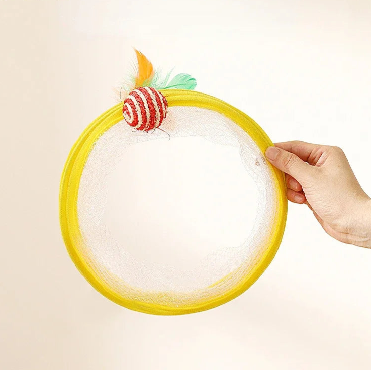 Hand holding a yellow folded spiral tunnel toy with a small ropeball on one end & a feather toy on the other end with  beige background,
